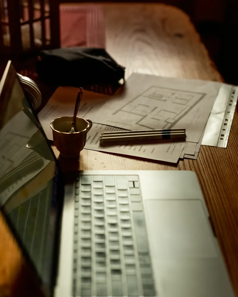 A laptop beside architectural floor plans and pencils on a warm wood desk