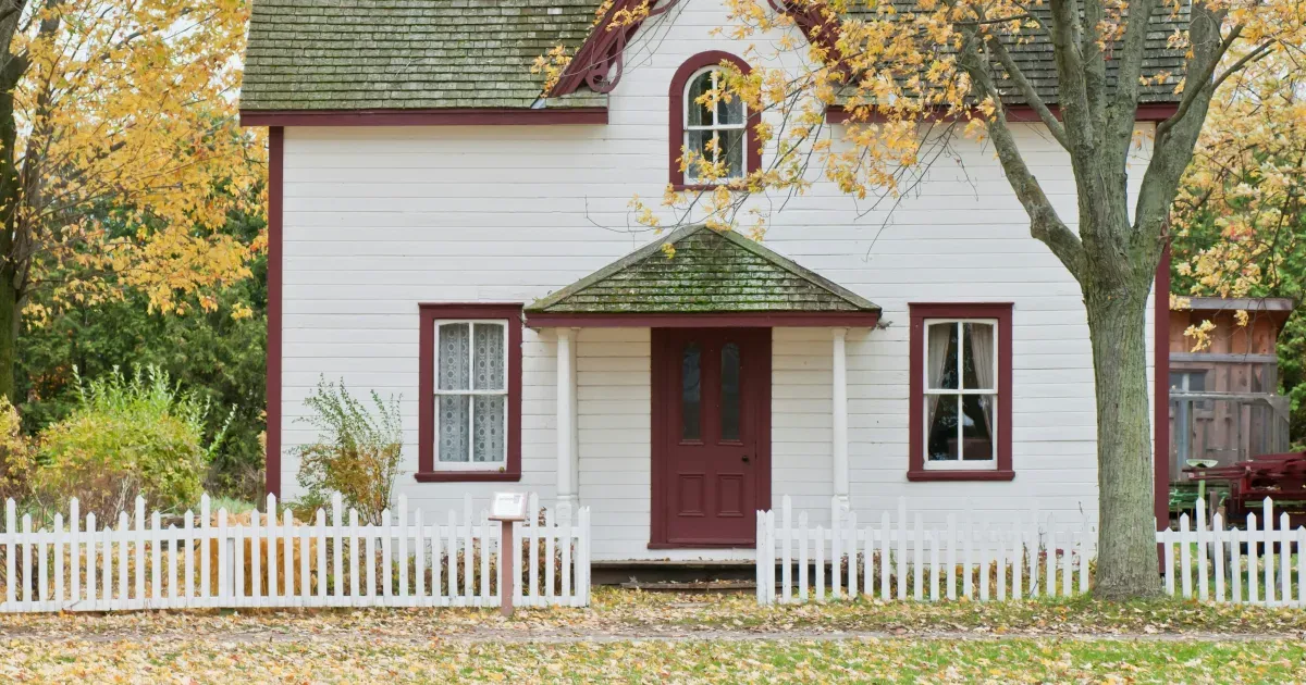 A white house with red trim and a white picket fence framed by autumn trees